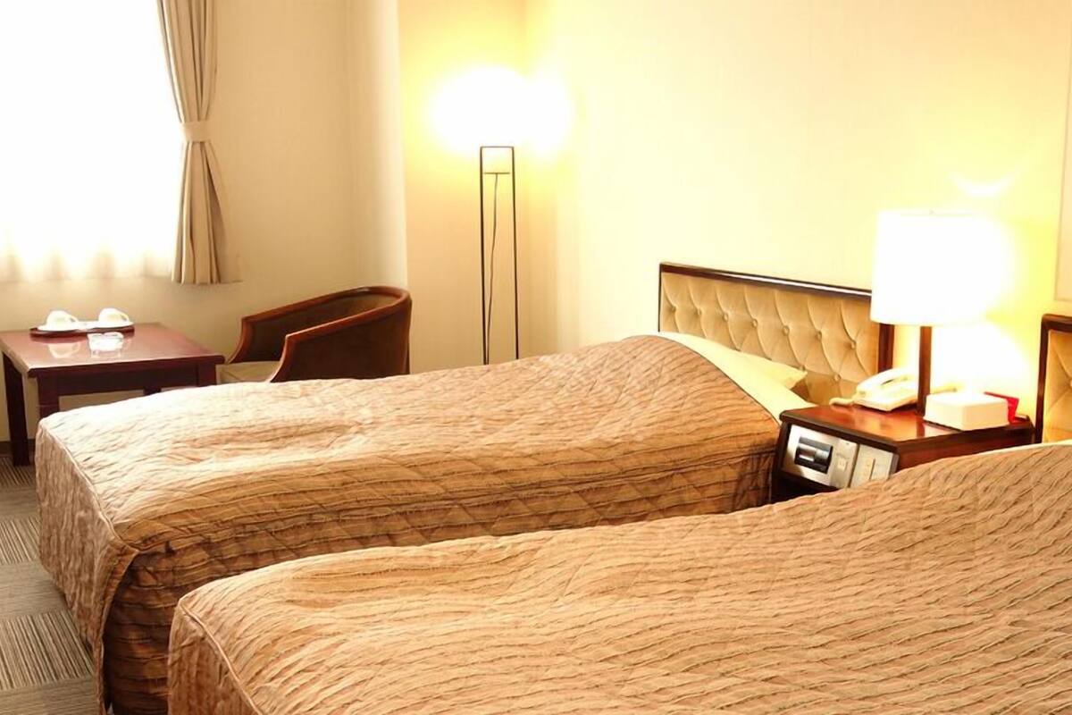 A hotel room with two beds, a desk, a chair, a lamp, and a bedside table.