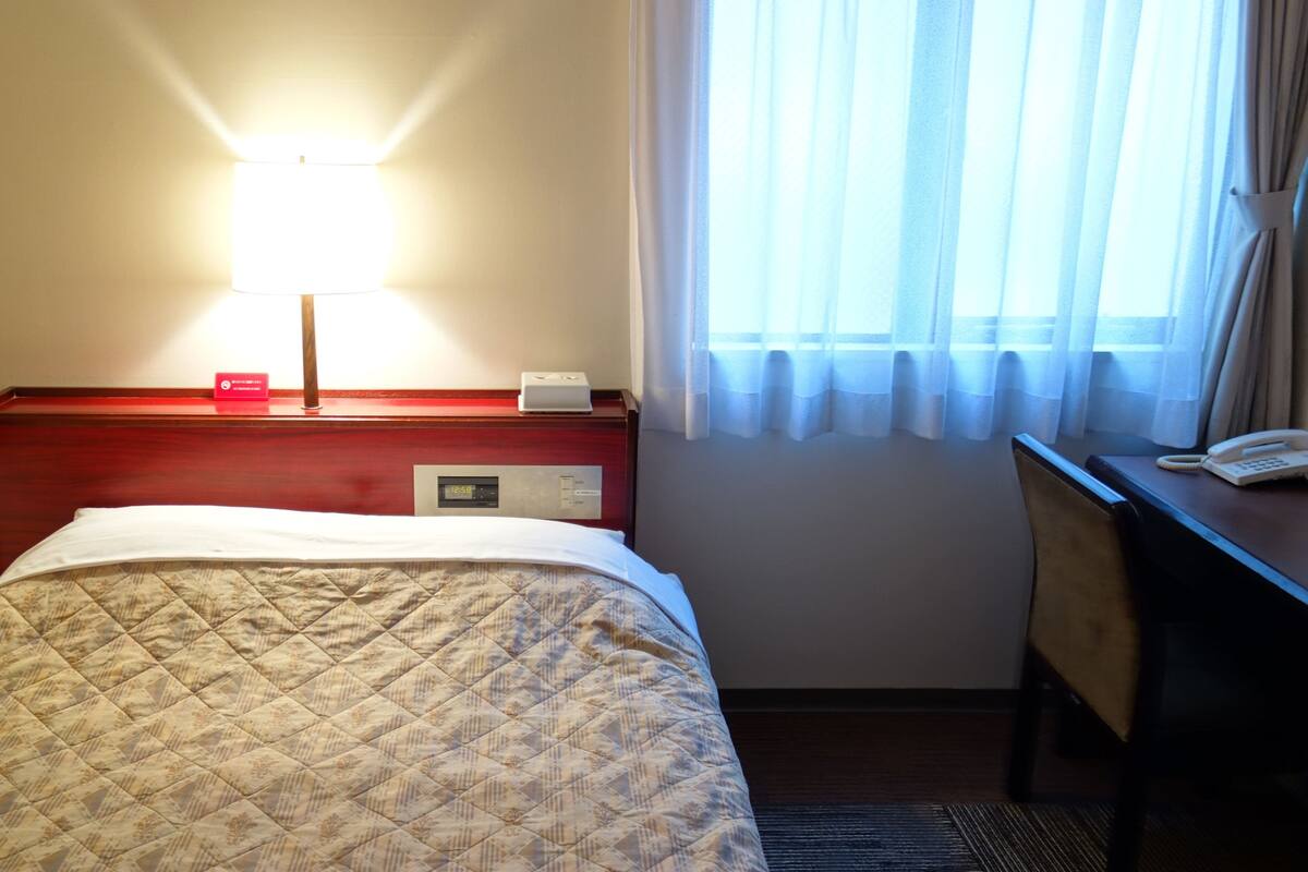 A hotel room with a bed, a bedside table with a lamp, a digital clock, and a telephone.