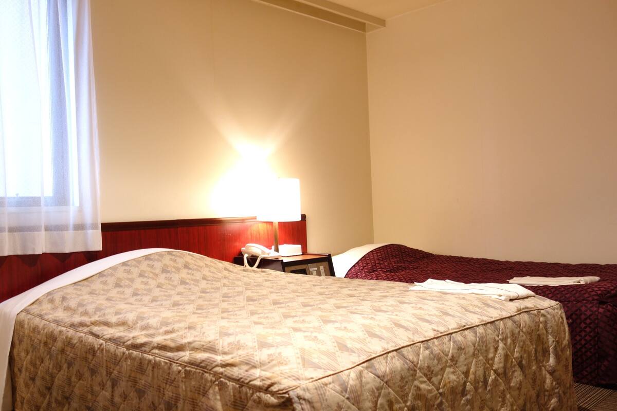 A hotel room with two beds, a bedside table with a lamp, and a window with sheer curtains.
