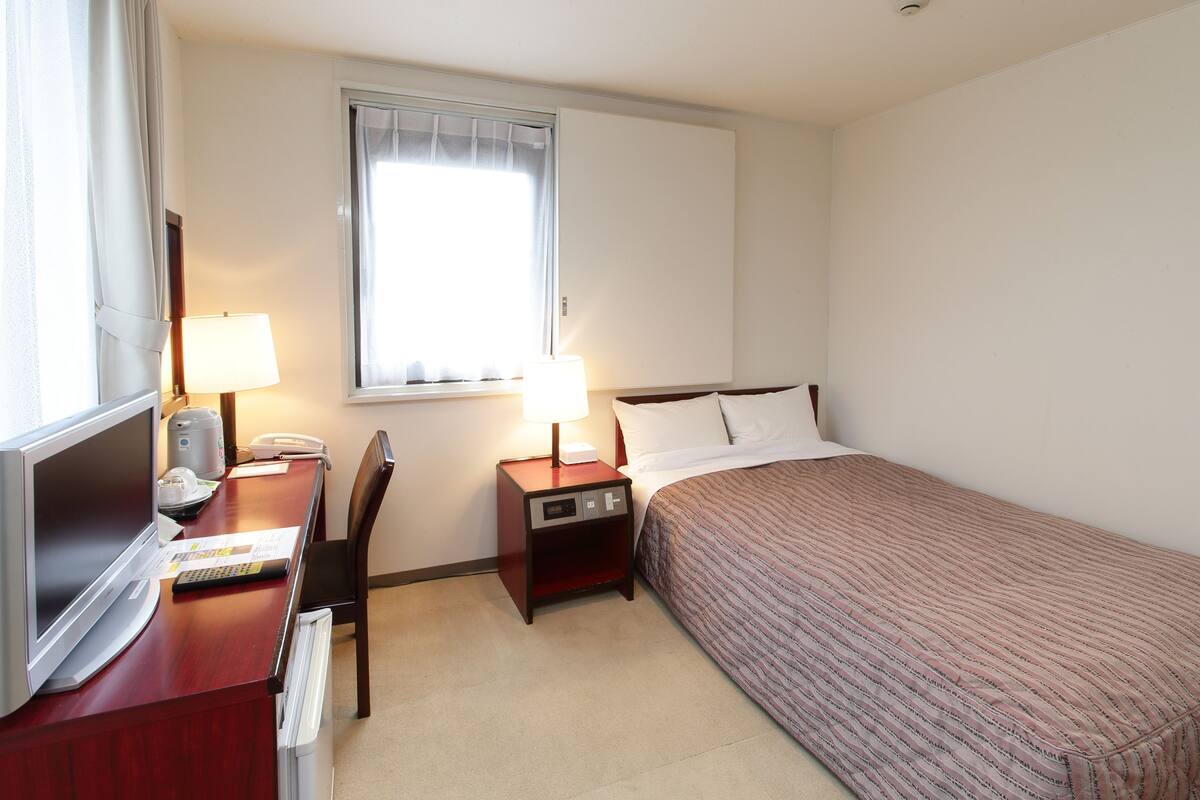 A hotel room with a bed, a desk with a lamp, a TV, and a bedside table with a phone and a clock.
