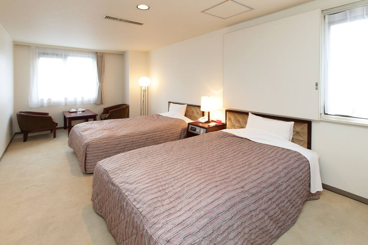A hotel room with two beds, a desk, a chair, a lamp, and a window.