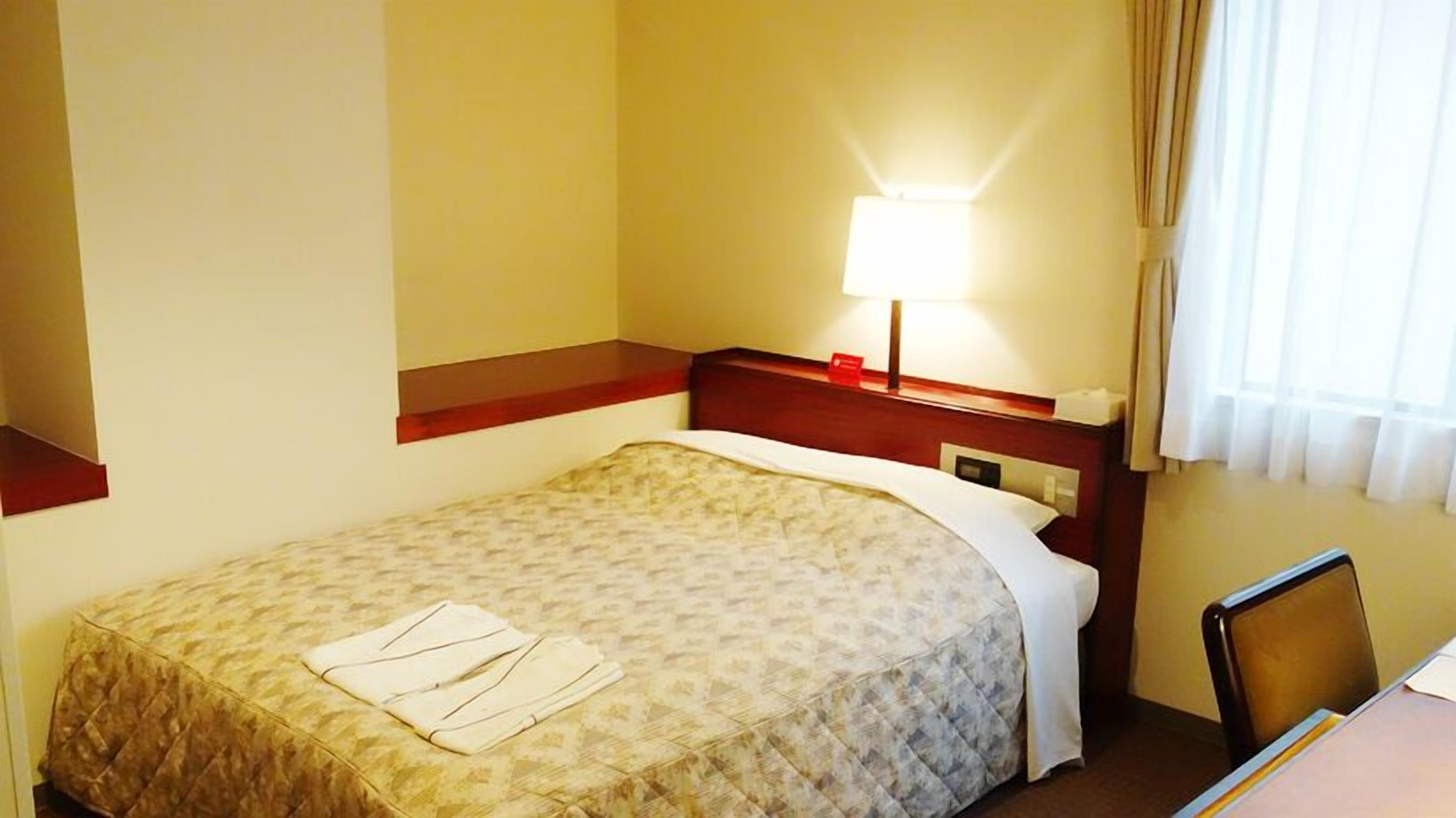 A hotel room with a bed, a desk, a chair, a lamp, and a window with curtains.