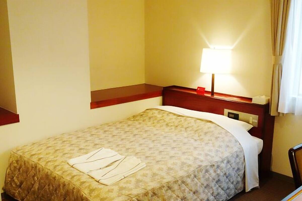 A hotel room with a bed, a desk, a chair, a lamp, and a window with curtains.