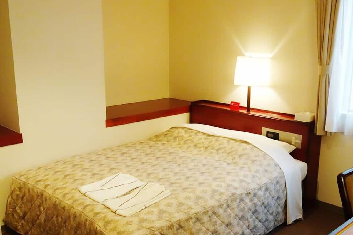 Quarto de hotel com cama, escrivaninha, cadeira, luminária e janela com cortinas.