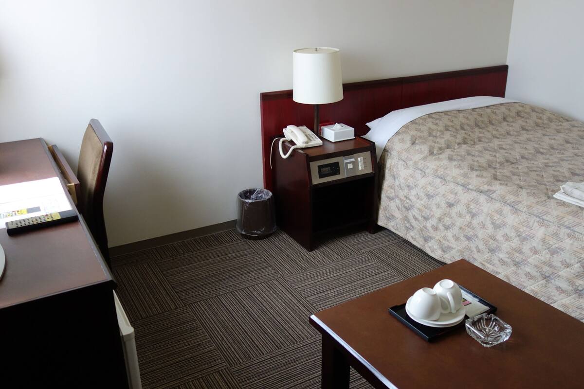 A hotel room with a bed, a desk with a lamp, a trash bin, and a tray with cups and a glass.