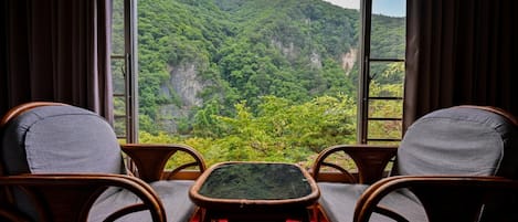 A room with two wicker chairs and a small table, overlooking a lush green valley with mountains.