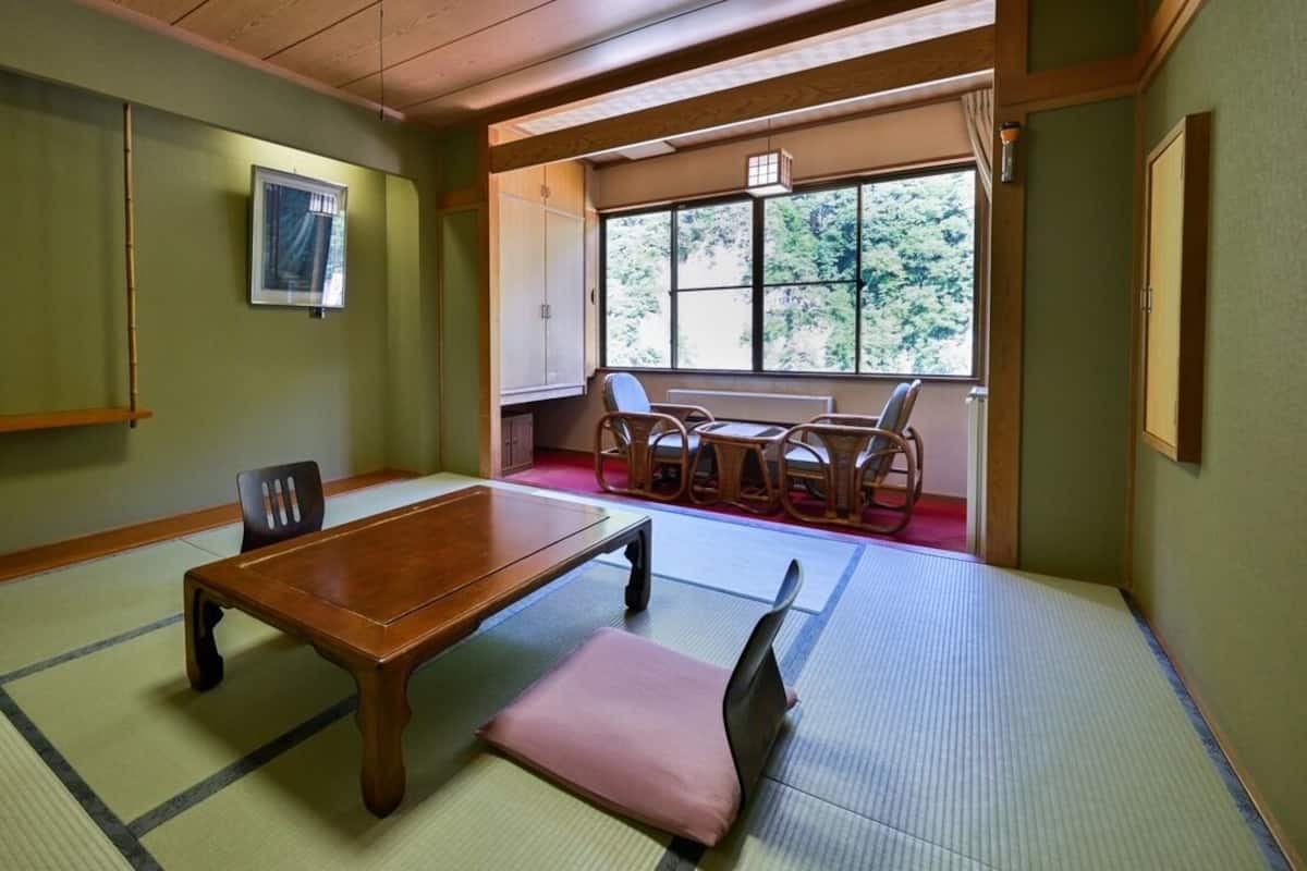 A traditional tatami room with a wooden table, tatami mats, a window with a view of trees, and a low wooden ceiling.