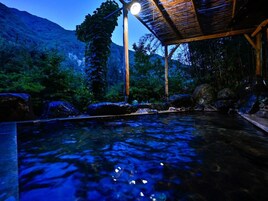 A tranquil outdoor hot spring area with a wooden structure overhead, surrounded by lush greenery and a mountainous backdrop.