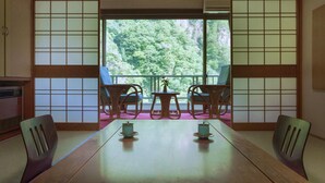 Une pièce de style japonais traditionnel, avec un sol recouvert de tatamis, une petite table et deux chaises, et une vue sur une zone boisée à travers un paravent shoji.