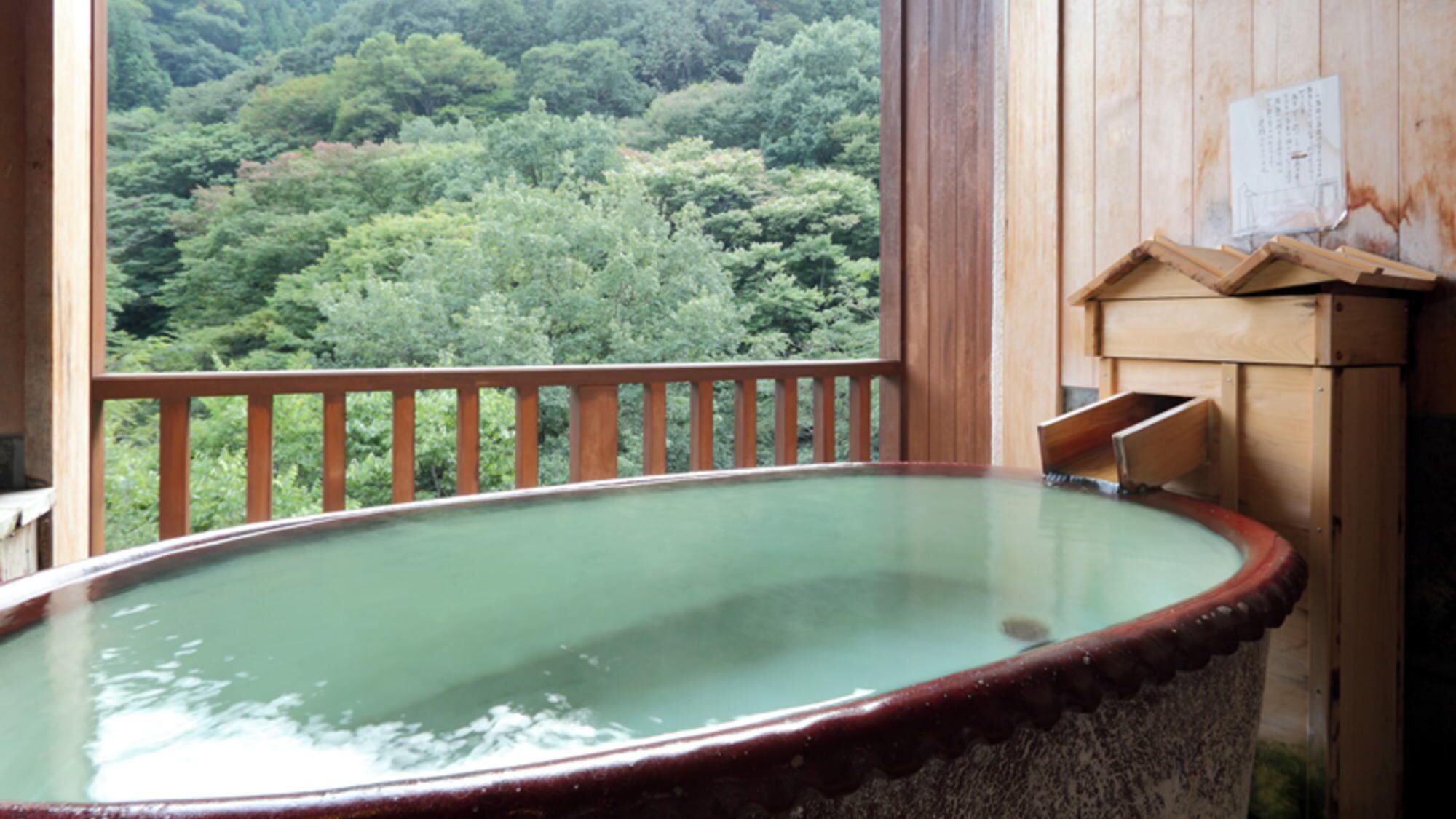 Un baño de aguas termales con barandilla de madera, un armario de madera y vistas a un área boscosa.