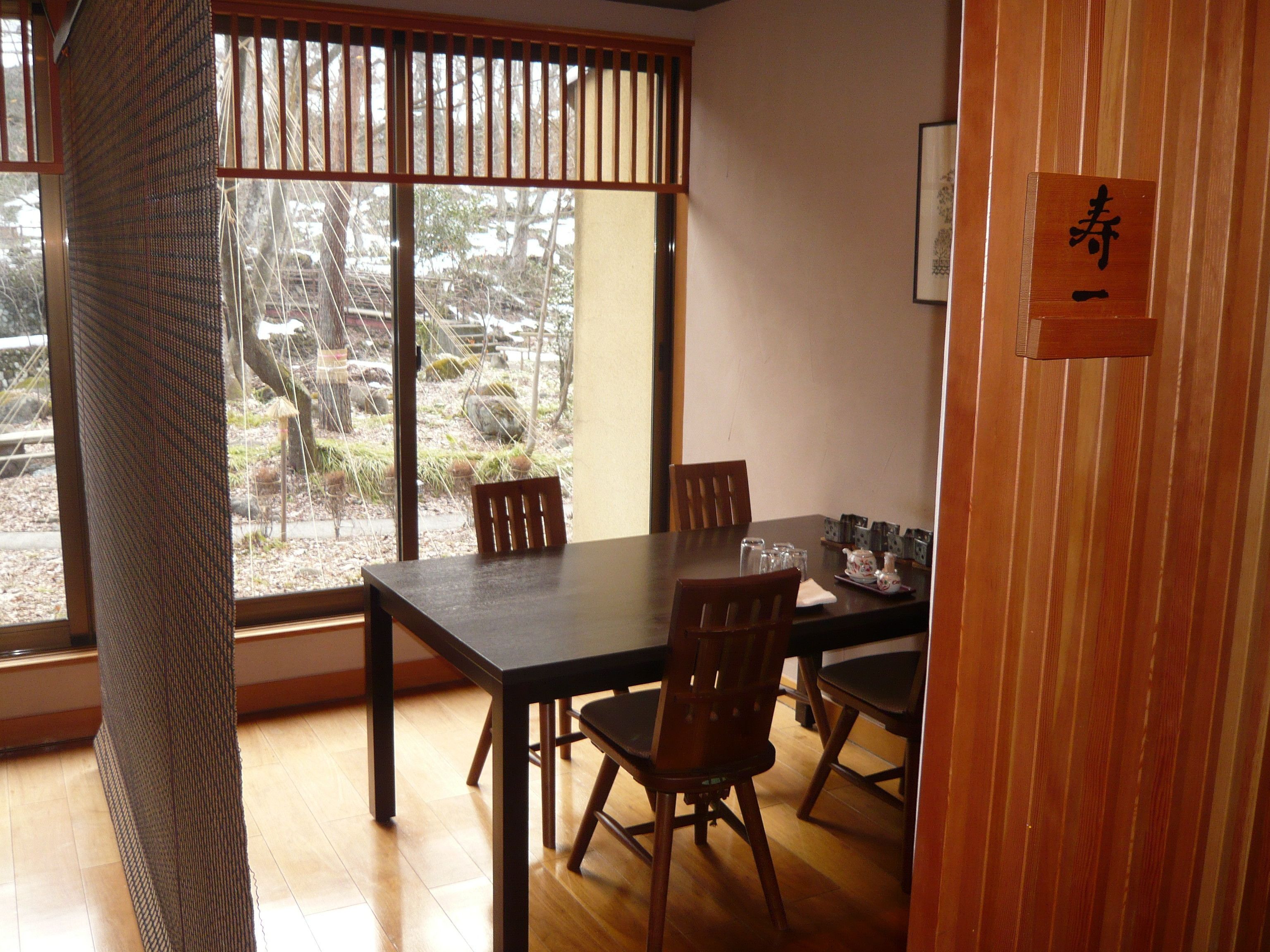 Een eethoek met een houten tafel en stoelen, een uitzicht door een shoji-scherm, en een bord met Japanse tekens.