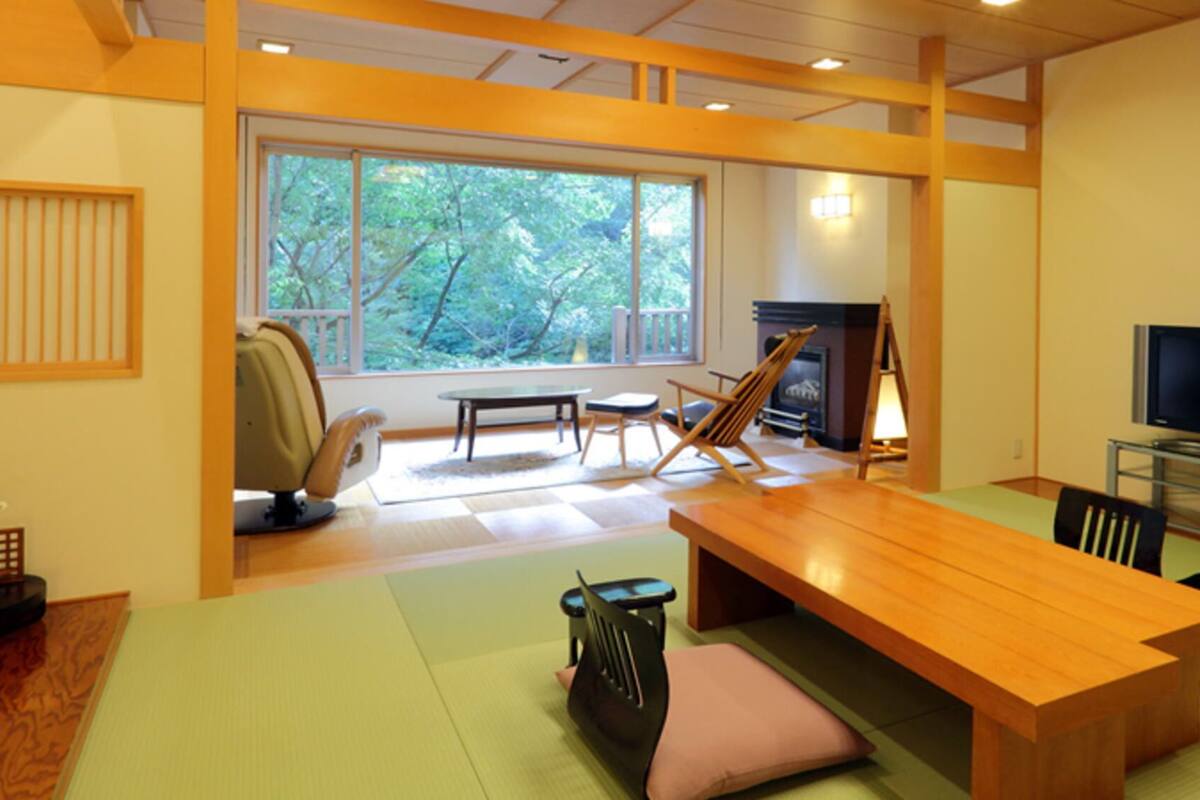 Una sala de estar moderna con una mesa de madera, sillas, un sofá, una chimenea, un televisor y ventanas amplias con vistas a árboles.