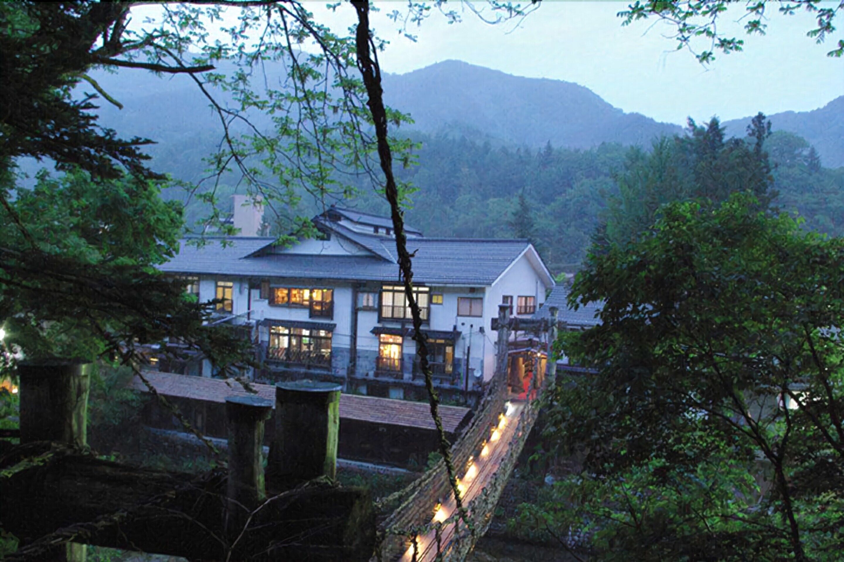 A traditional Japanese house with a wooden deck and string lights, surrounded by lush greenery and mountains in the background.