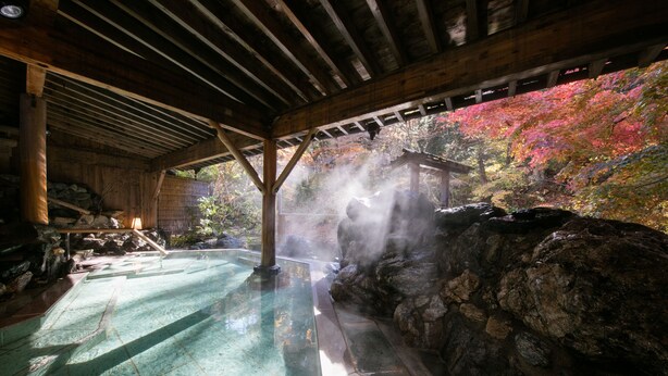 Un onsen japonés tradicional con una bañera de piedra, vigas de madera y vistas a follaje otoñal colorido.