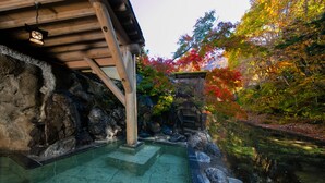Un onsen japonés tradicional con escalones de piedra que conducen a una piscina cristalina, rodeado de follaje otoñal y una estructura de madera.
