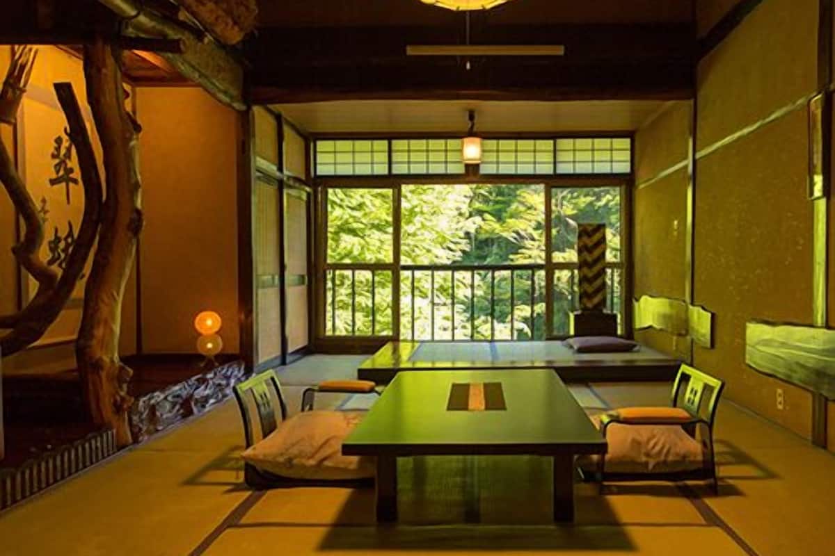 Um quarto tradicional japonês com piso de tatame, uma mesa baixa e vista para o verde através de janelas amplas.