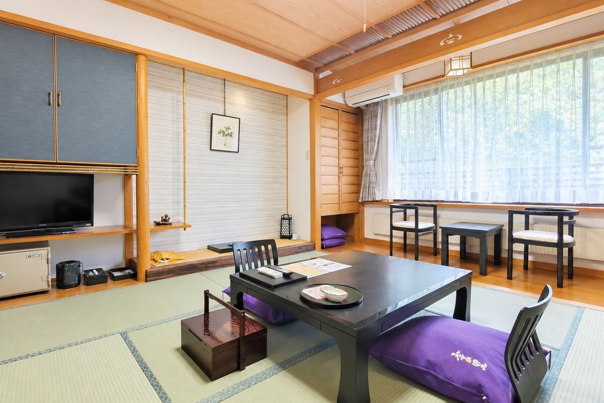 Une pièce de style japonais traditionnel, avec un sol recouvert de tatamis, une télévision, un meuble et une fenêtre avec des rideaux.