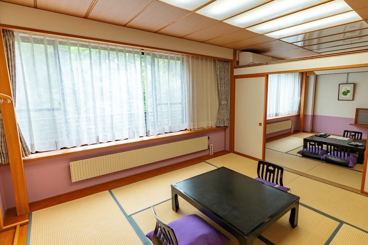 Een kamer met tatami vloerbedekking, een zwarte tafel, paarse kussens, een radiator en een raam met dunne gordijnen.