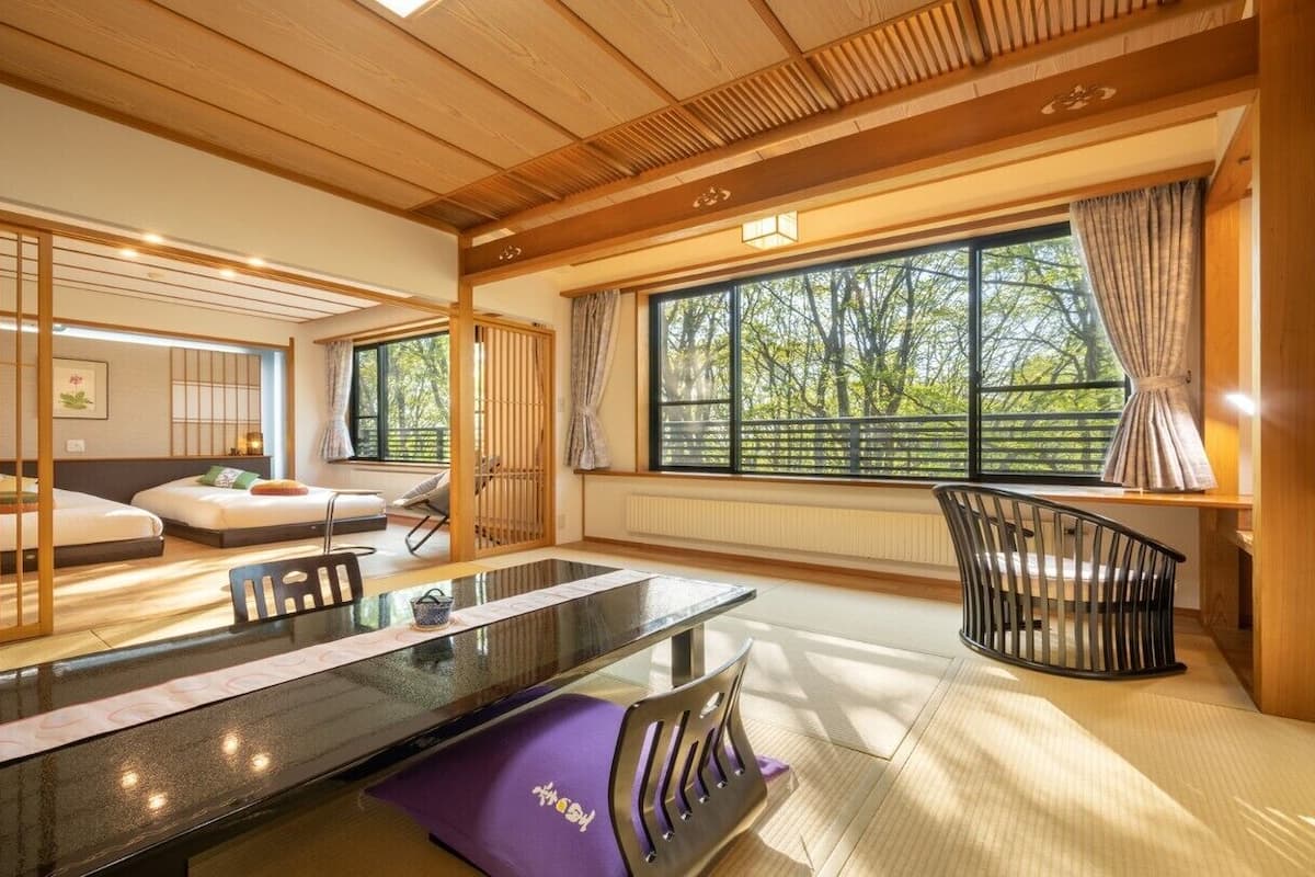 Een traditionele Japanse kamer met tatami vloerbedekking, een lage tafel en uitzicht op bomen door grote ramen.