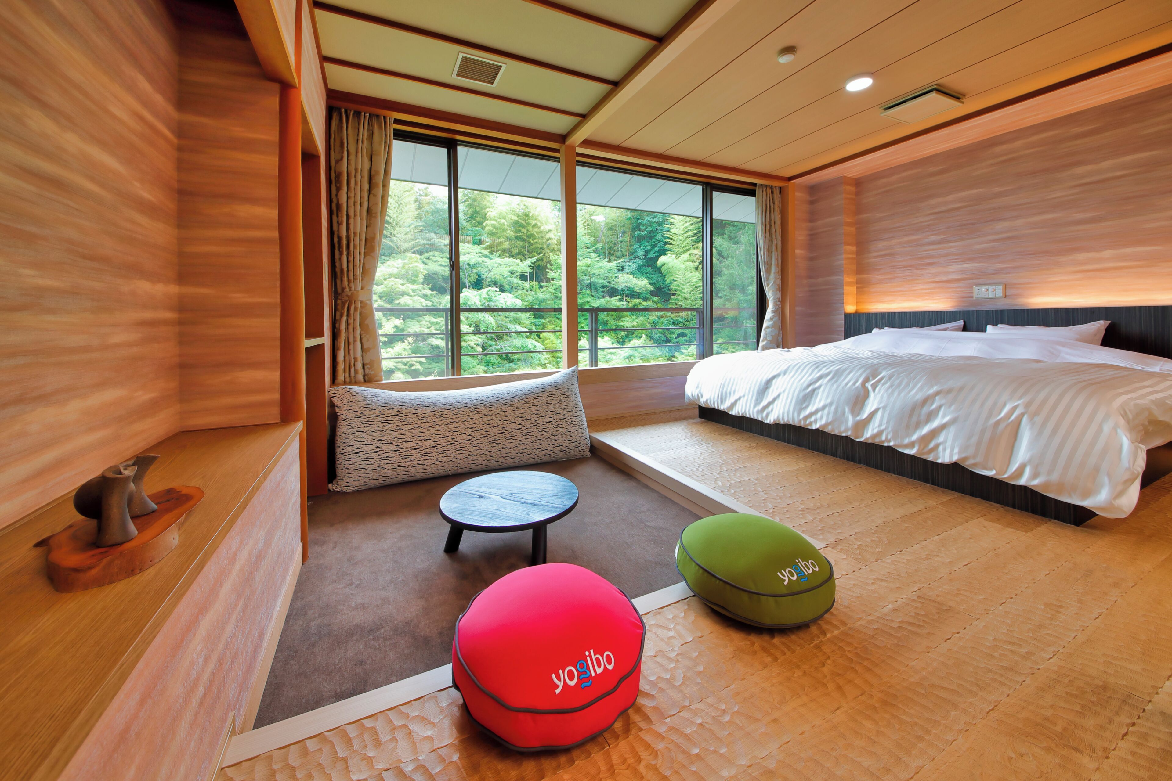 A modern hotel room with a large bed, a seating area with a round table, and a view of the forest through large windows.