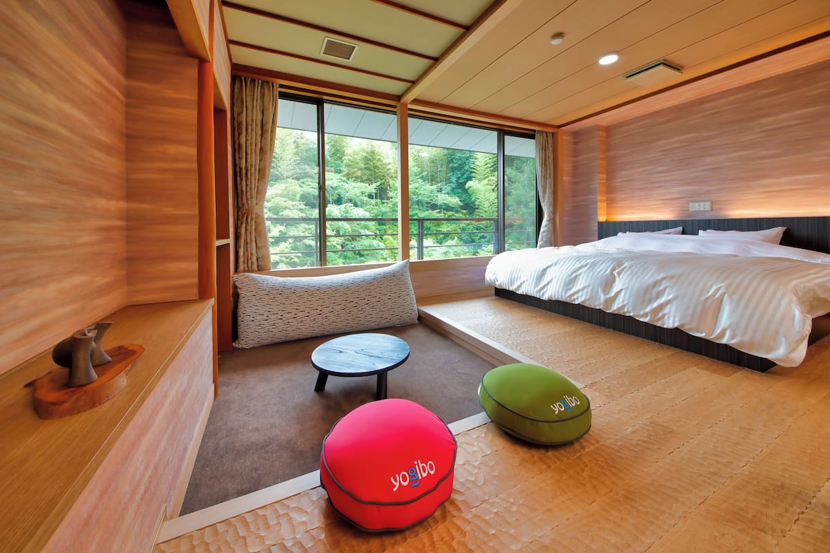 A modern hotel room with a large bed, a seating area with a round table, and a view of the forest through large windows.