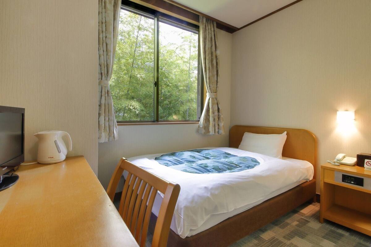 A hotel room with a bed, a desk with a television, a kettle, and a nightstand with a phone and a sign.