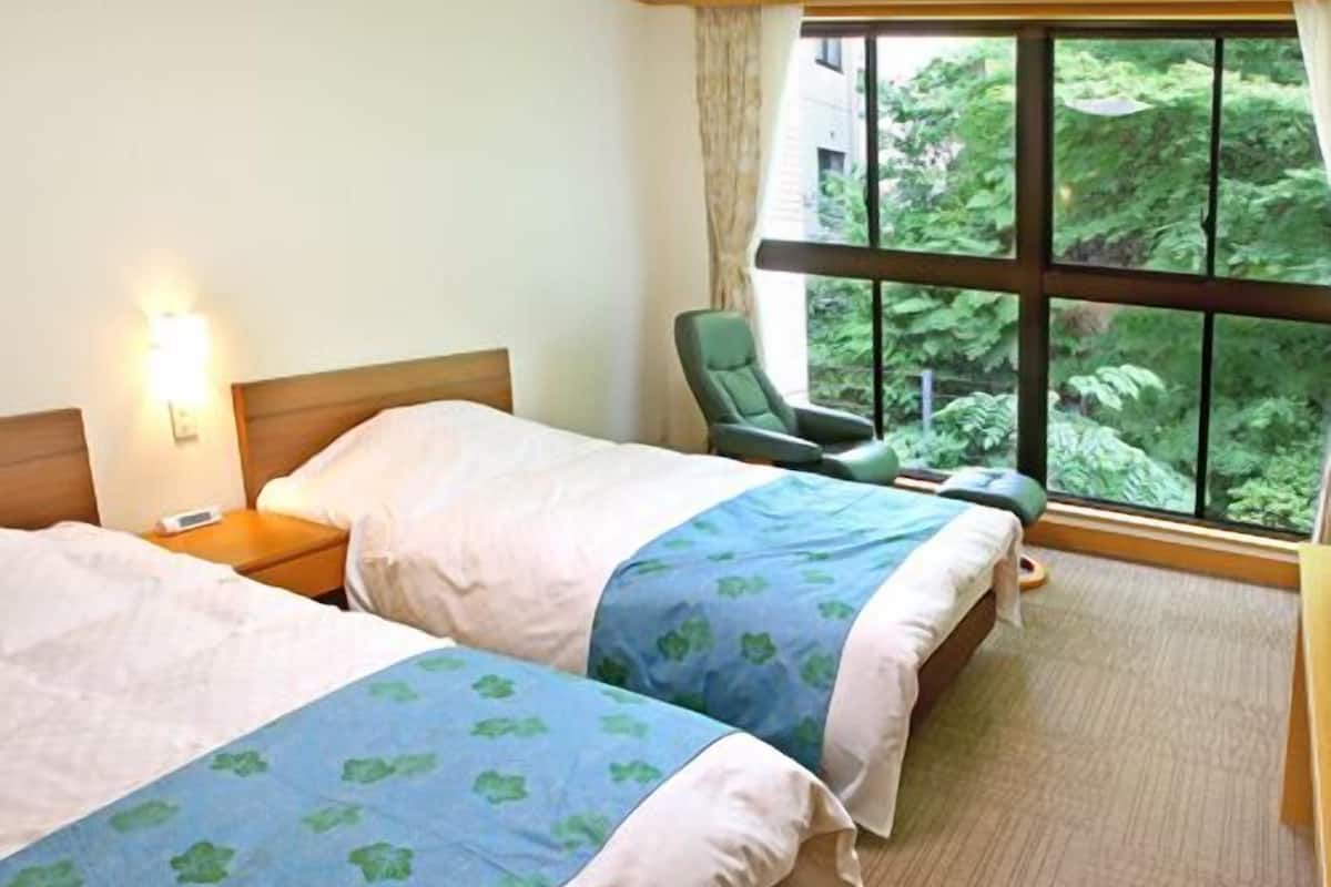 A hotel room with two beds, a chair, a nightstand, and a large window with a view of greenery.