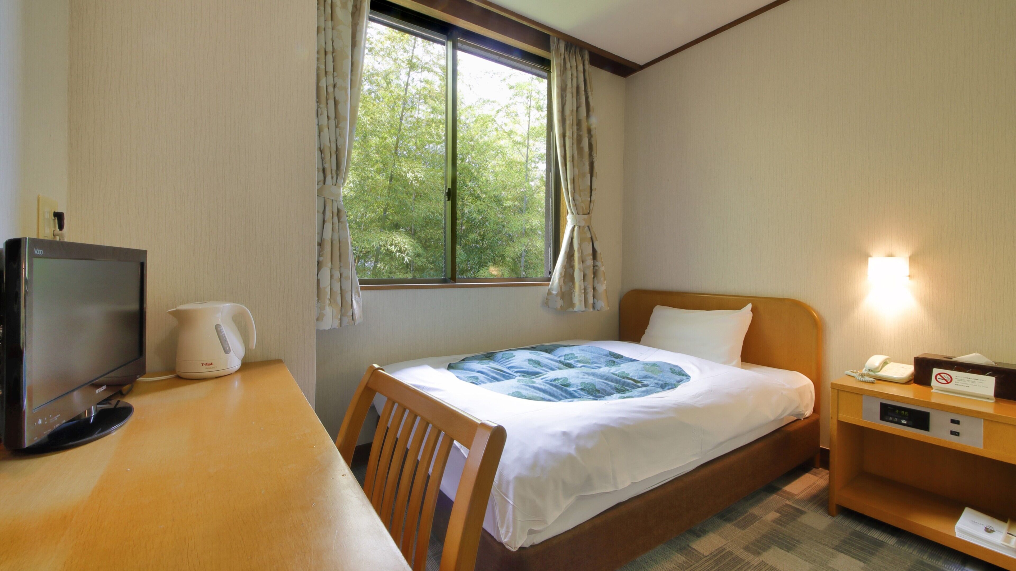 A hotel room with a bed, a desk with a computer, a kettle, and a nightstand with a phone.