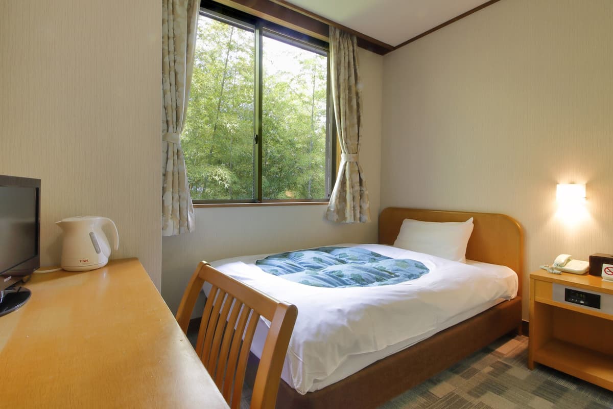 A hotel room with a bed, a desk with a computer, a kettle, and a nightstand with a phone.