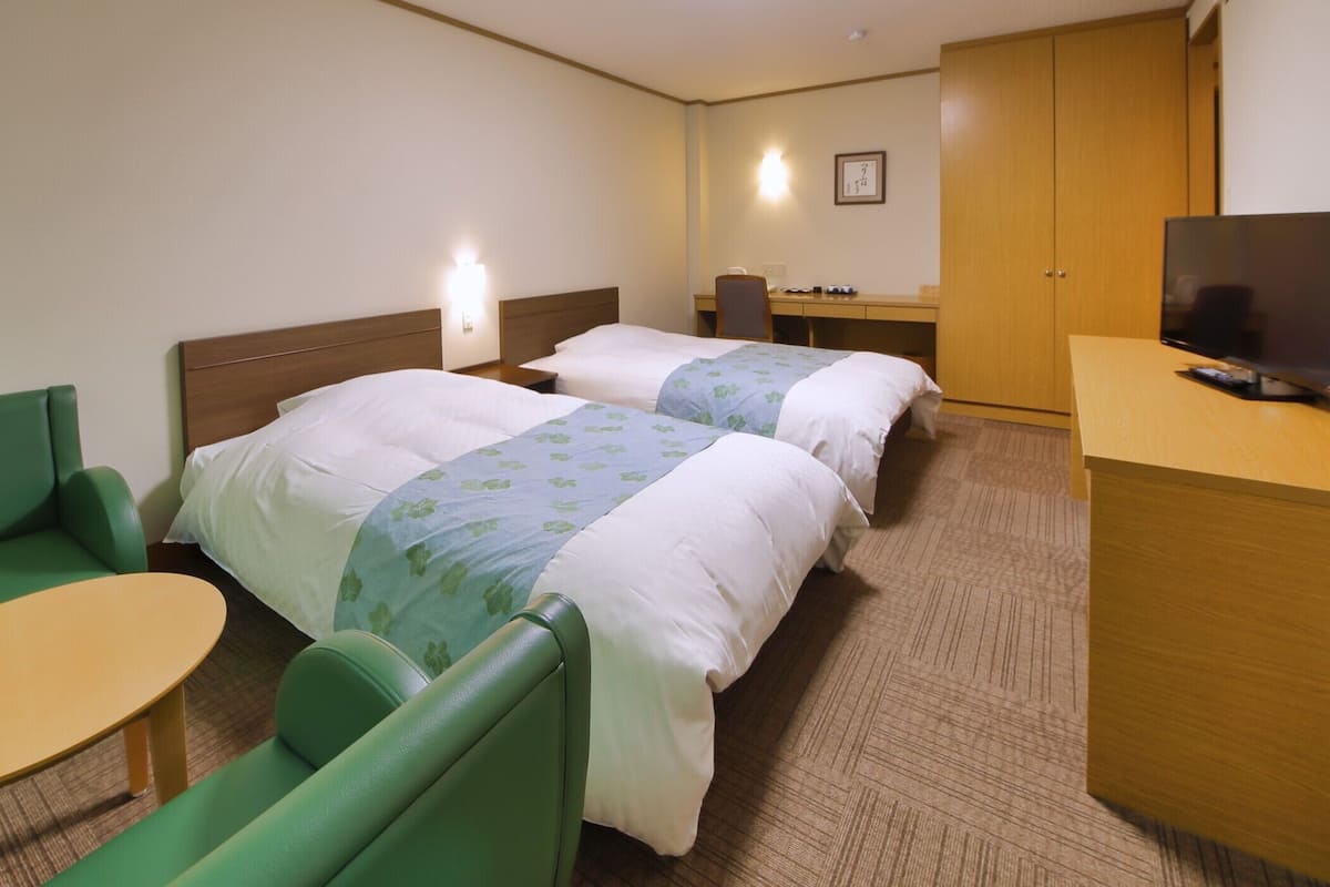 A hotel room with two beds, a green sofa, a small table, a TV, and a wooden wardrobe.