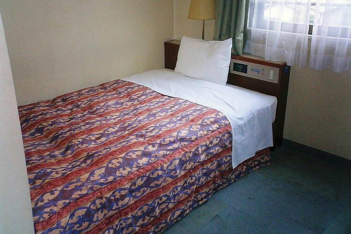 Una habitación de hotel pequeña y sencilla con una cama individual, una mesita de noche, una lámpara y una ventana con cortinas translúcidas.