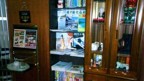 A room with a wooden cabinet containing various items, a shelf with magazines, and a small table with a teapot and cups. - Heisei Hotel (Chikusei)