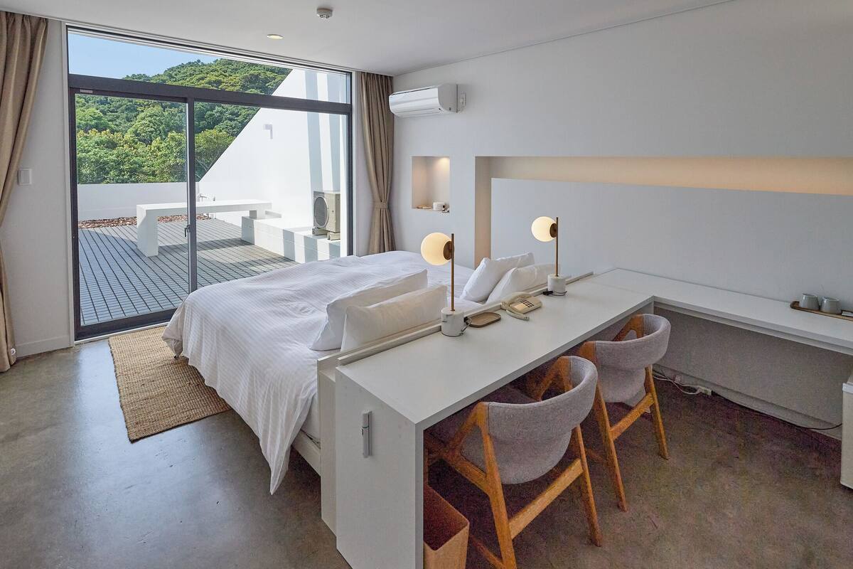 A modern hotel room with a bed, desk, chairs, and a view of greenery outside.