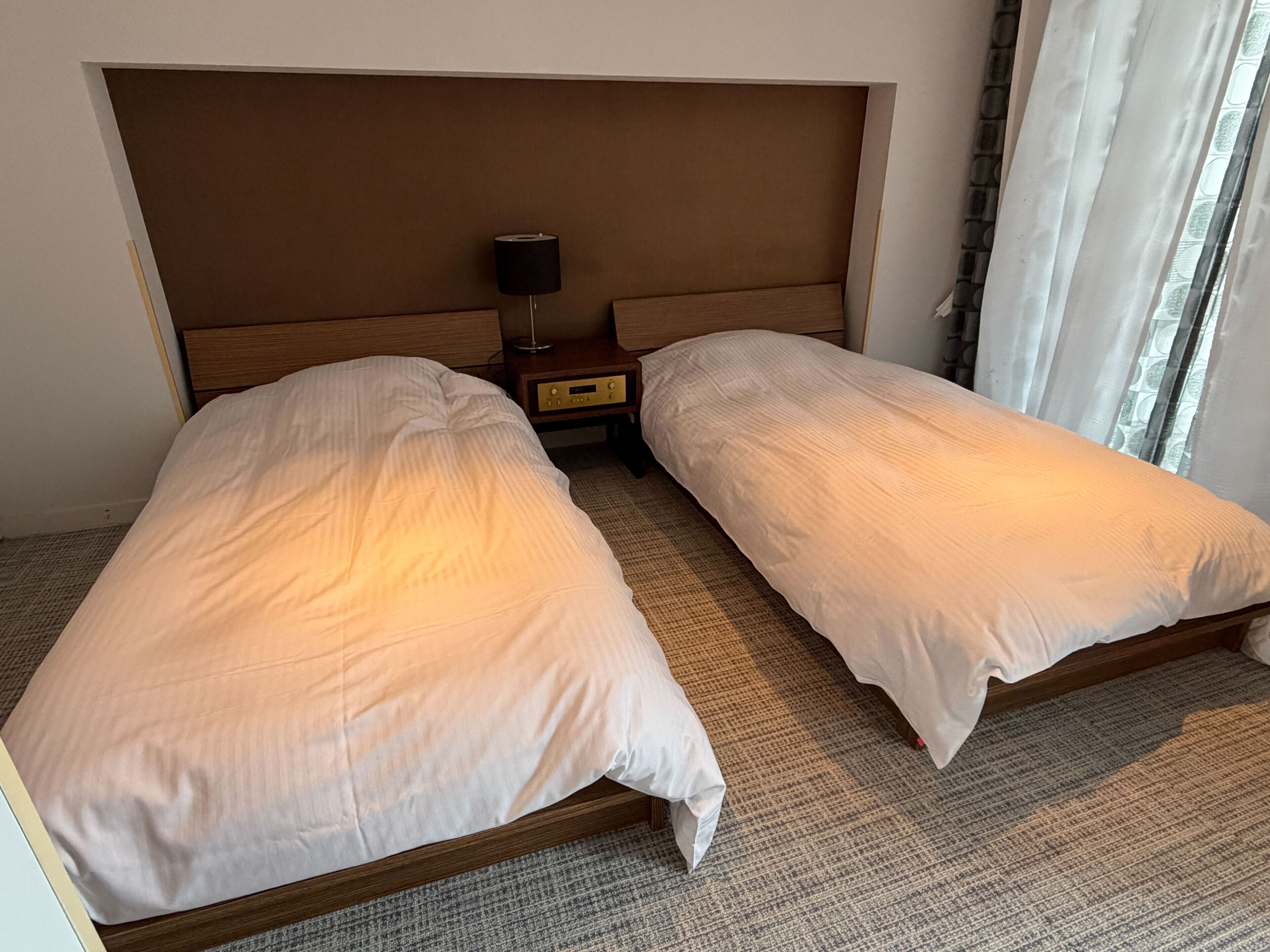 Hotellihuone, jossa on kaksi erillistä sänkyä, yöpöytä lampun kanssa ja ikkuna, jossa on verhot.