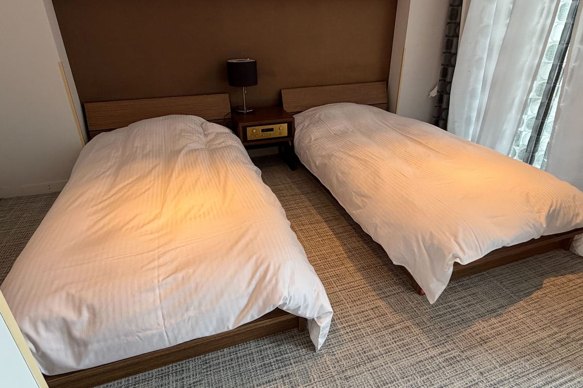 Hotellihuone, jossa on kaksi erillistä sänkyä, yöpöytä lampun kanssa ja ikkuna, jossa on verhot.