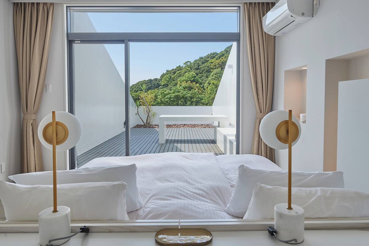 A hotel room with a large bed, two bedside tables, a view of a balcony with a table and chairs, and an air conditioning unit.