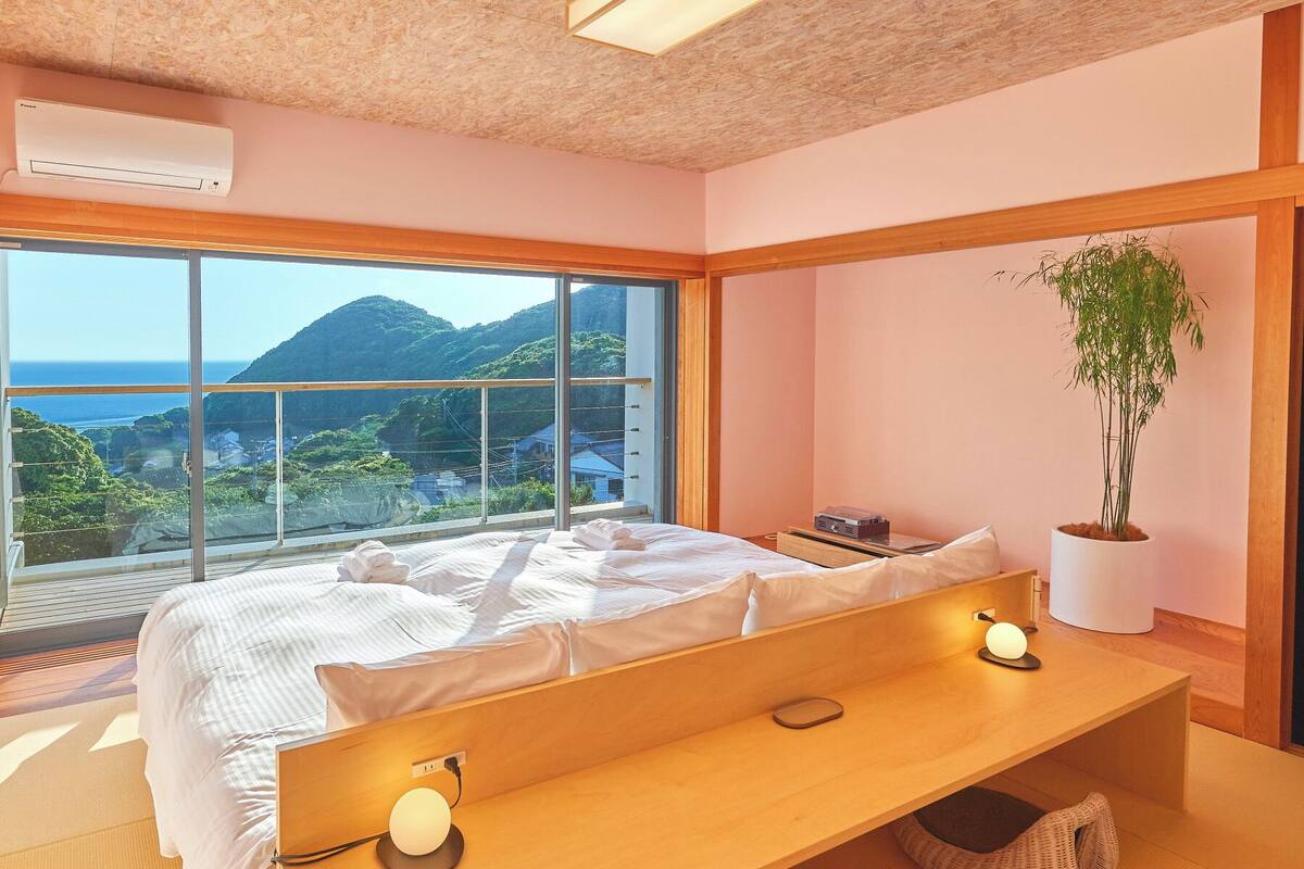 A bedroom with a large window, a bed, bedside lamps, a bedside table, a potted plant, and a view of the outdoors.