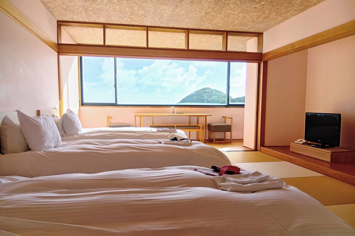 A hotel room with two beds, a desk, a chair, a TV, and a large window with a mountain view.