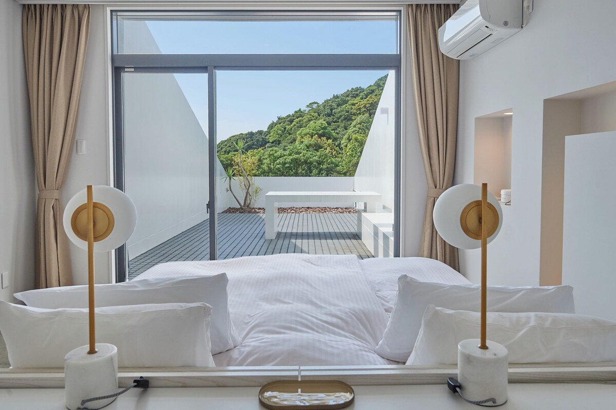 A hotel room with a large bed, two bedside tables, a view of a balcony with a table and chairs, and an air conditioning unit.