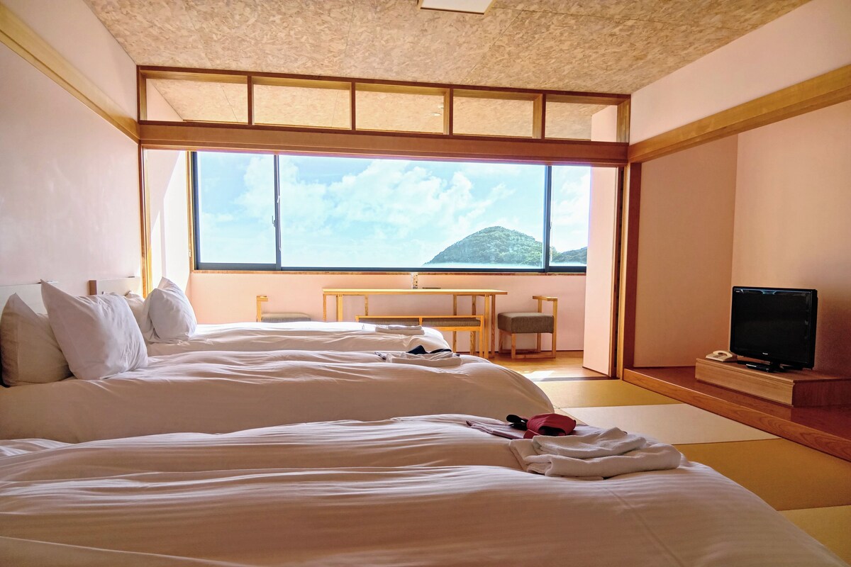 A hotel room with two beds, a desk, a chair, a TV, and a large window with a mountain view.