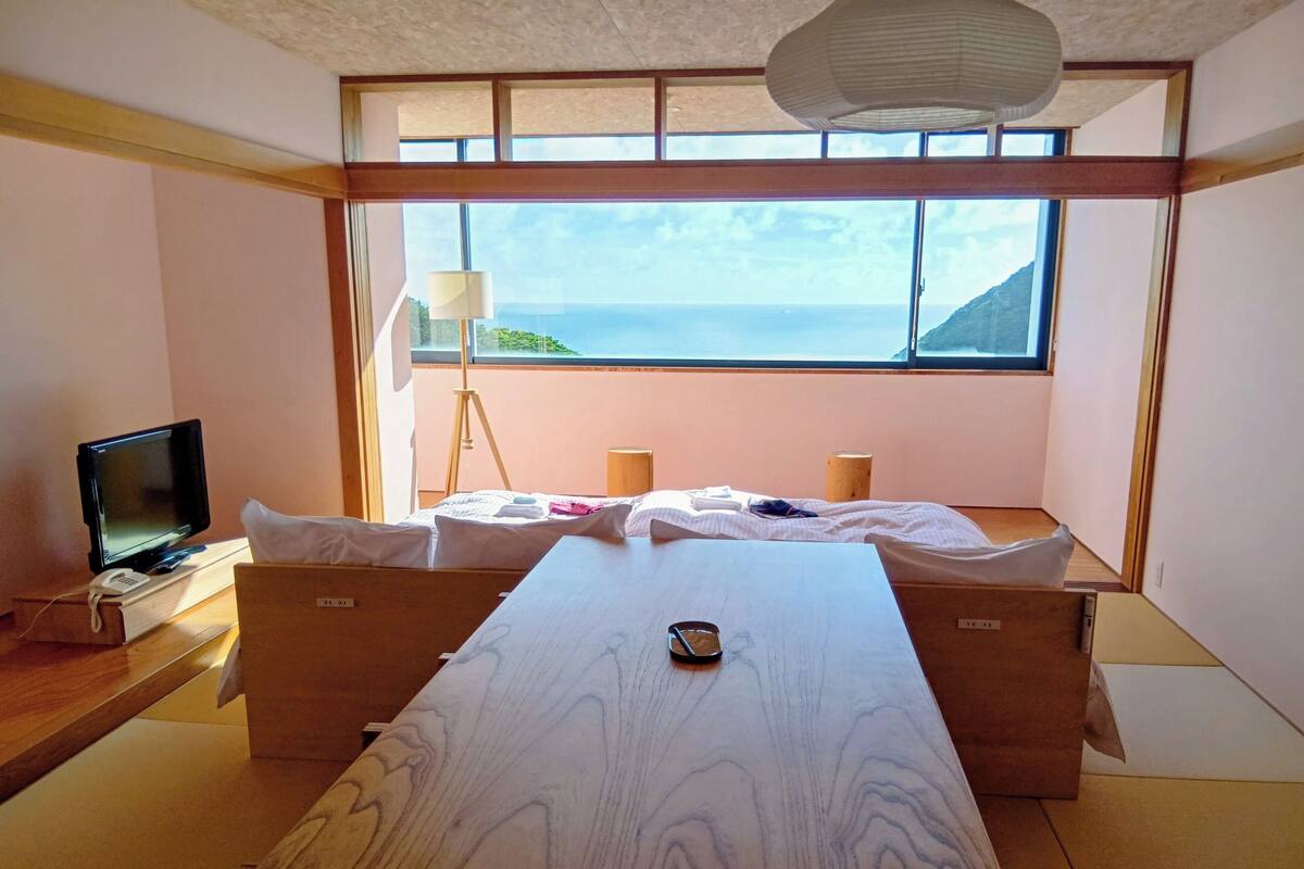 A room with a bed, a desk with a computer, a lamp, and a view of the sea.