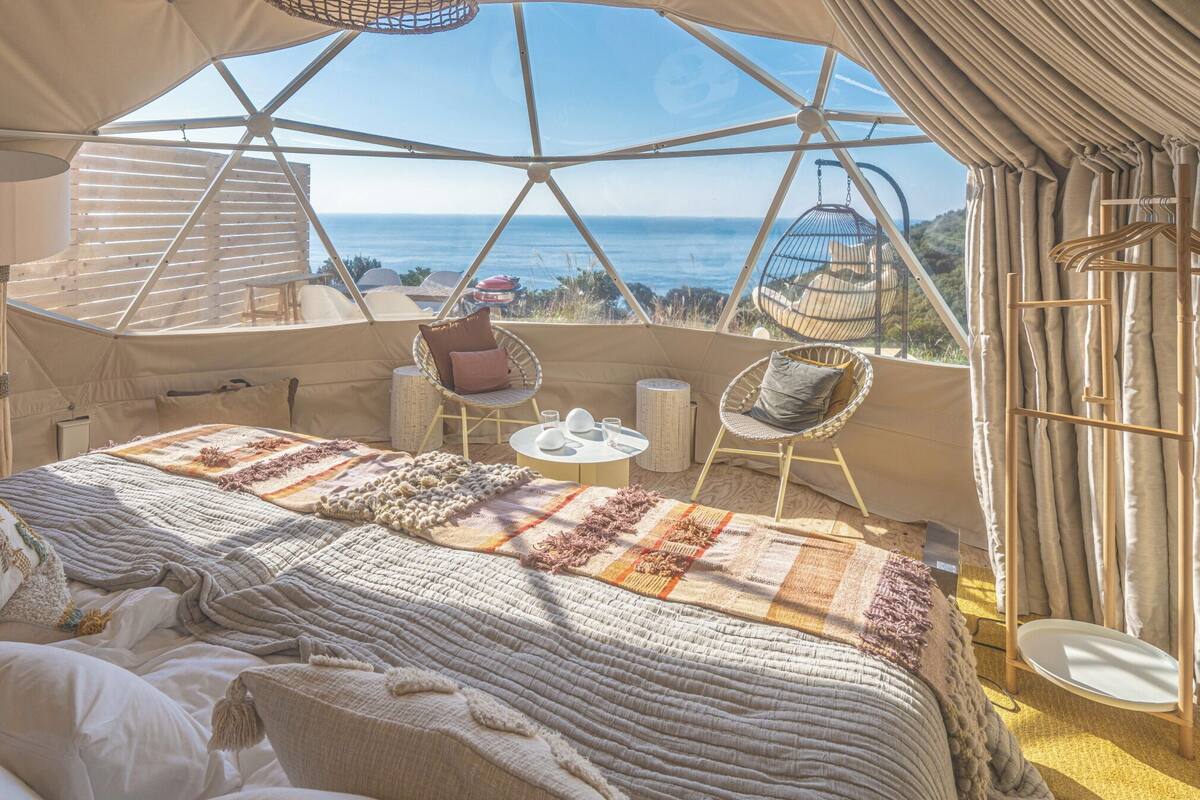 A tented accommodation with a bed, two chairs, a table, and a view of the sea.