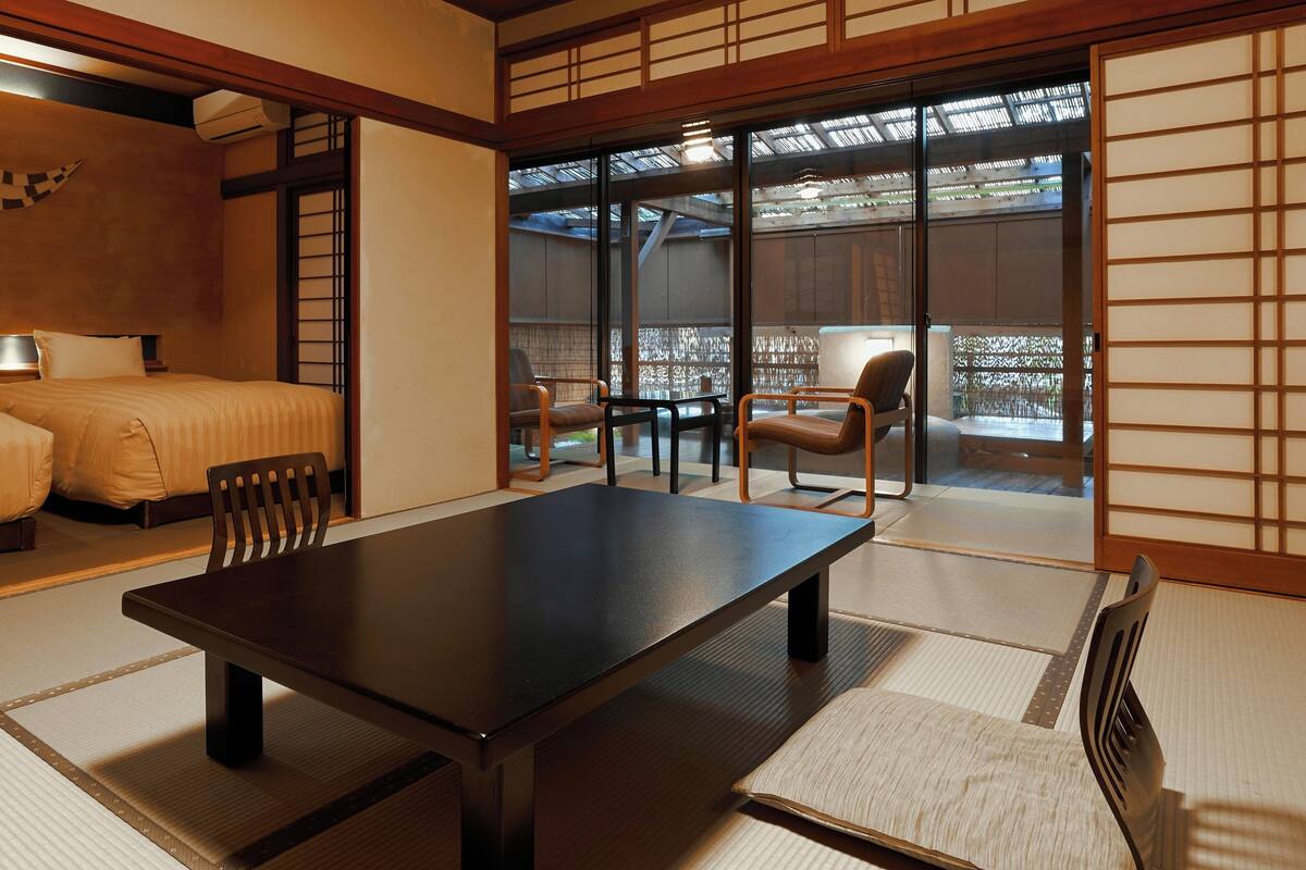 A traditional Japanese room with tatami flooring, a low table, a bed with a futon, and sliding doors leading to an outdoor area.