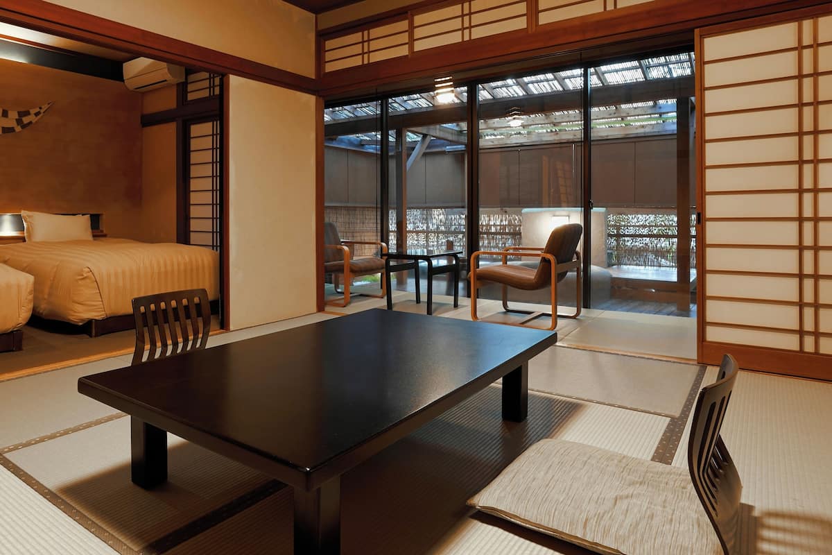 A traditional Japanese room with tatami flooring, a low table, a bed with a futon, and sliding doors leading to an outdoor area.
