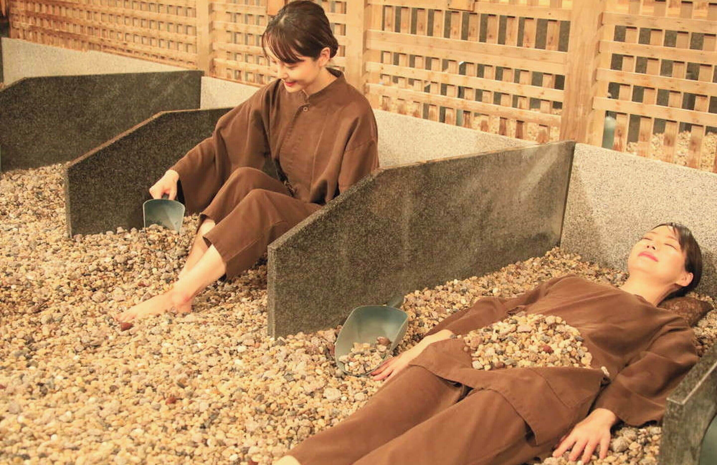 A spa area with two individuals in brown robes, one sitting and one lying down, surrounded by pebble-filled basins.
