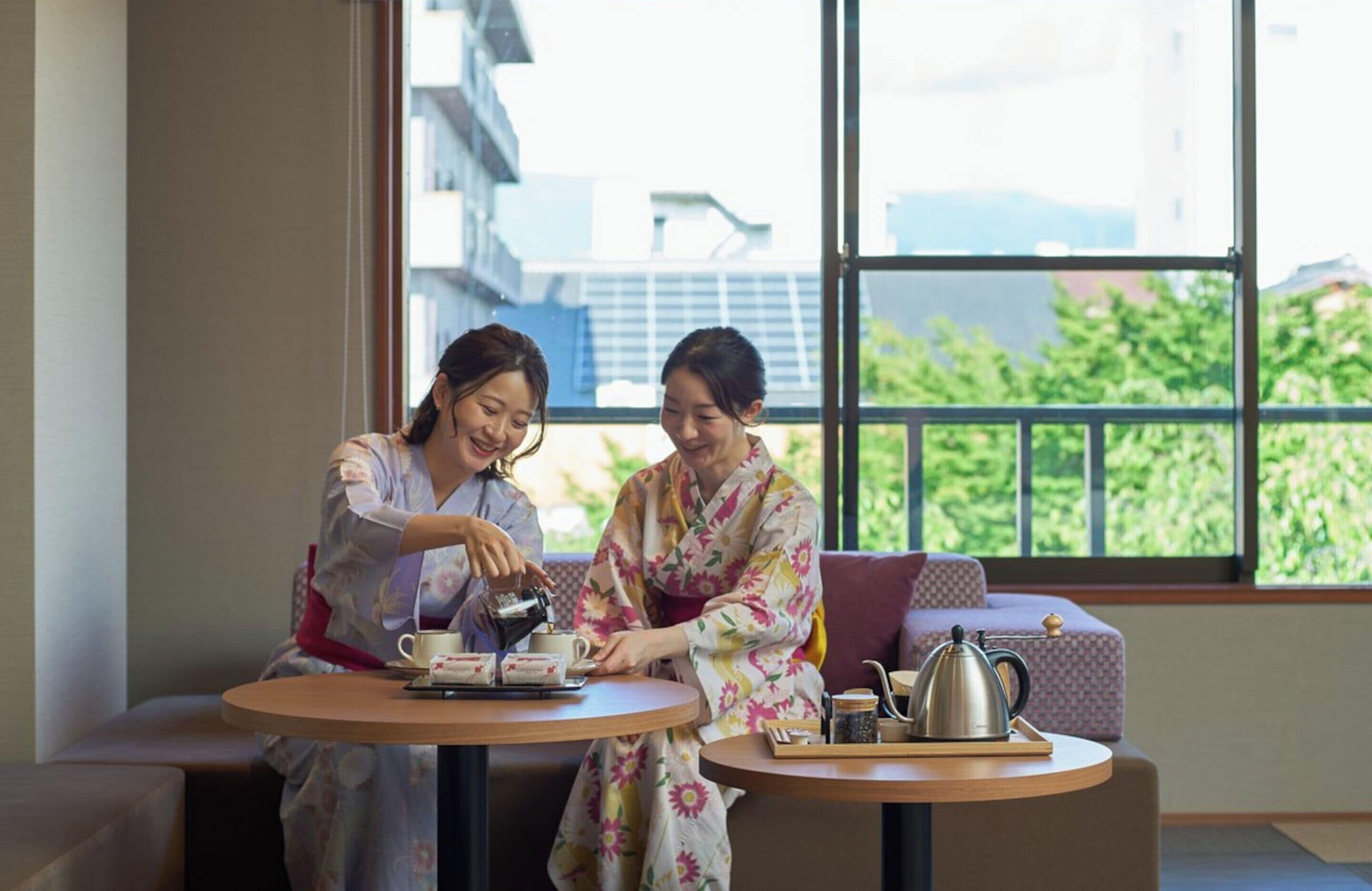 Two people in traditional attire at a round table with tea set, in a room with large windows and a view of buildings and greenery.