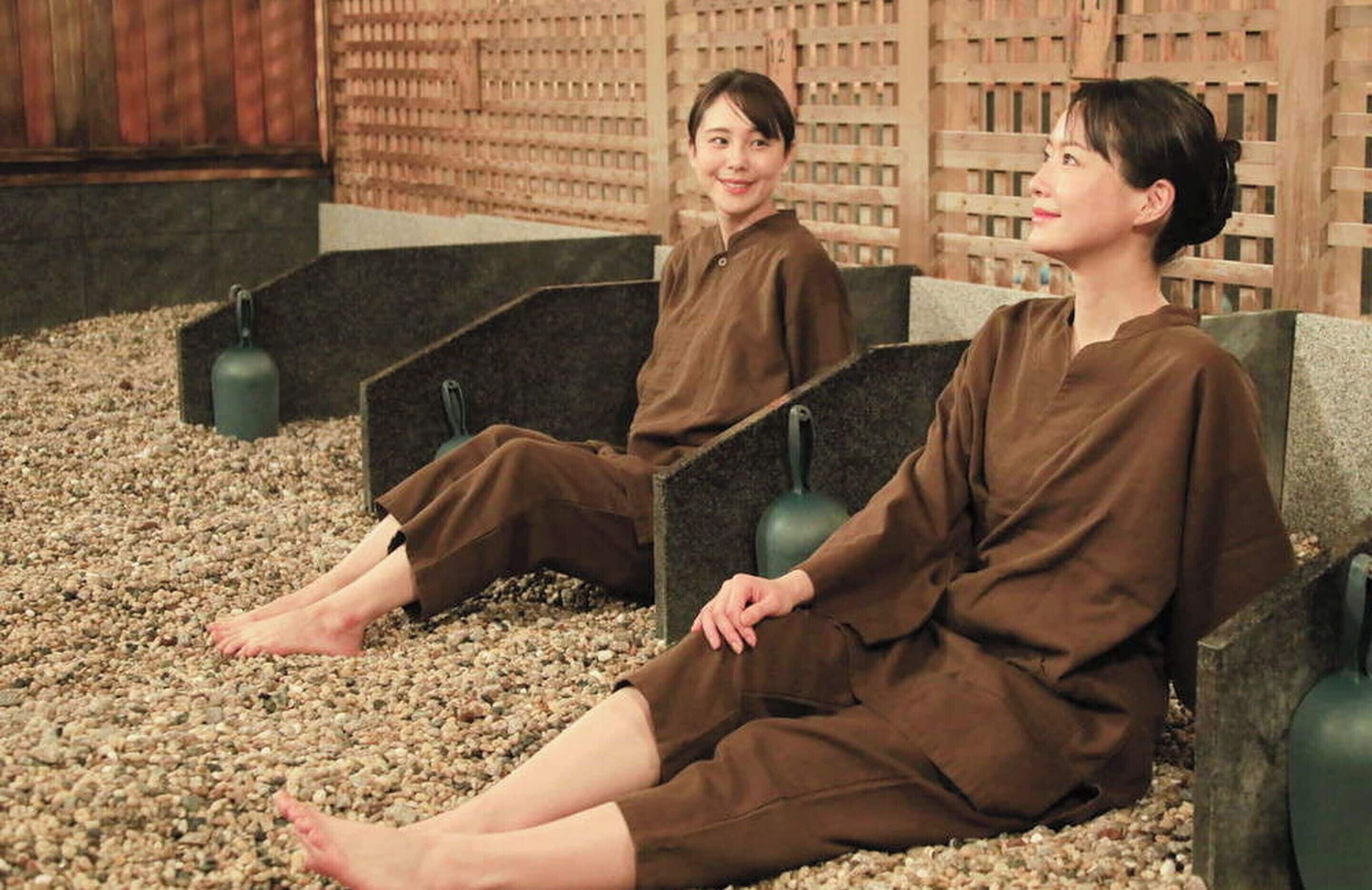 Two people sitting on benches in a room with a wooden lattice wall and a pebble floor.