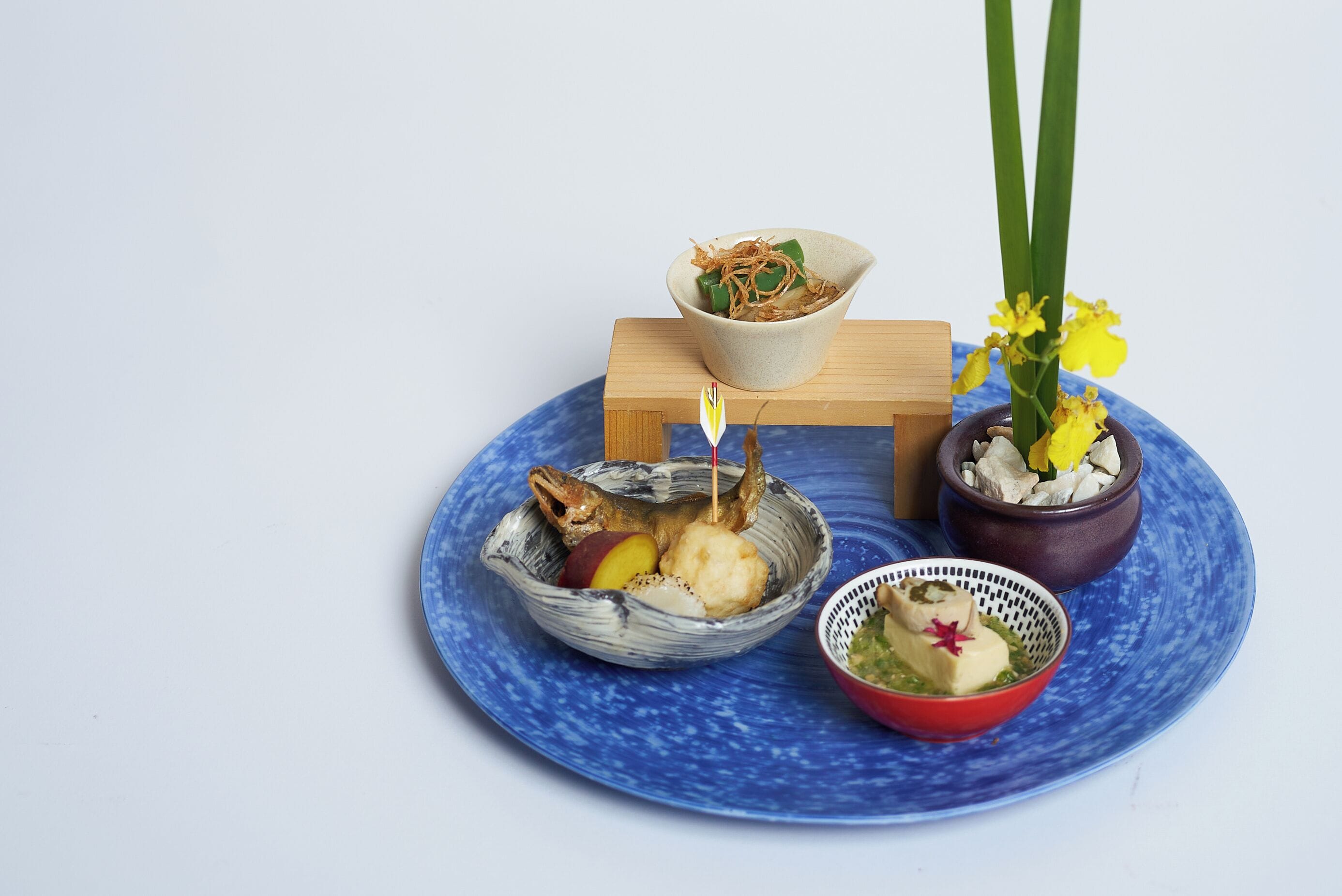 Un plato azul con diversos alimentos, un bloque de madera pequeño y una planta en maceta con flores amarillas.