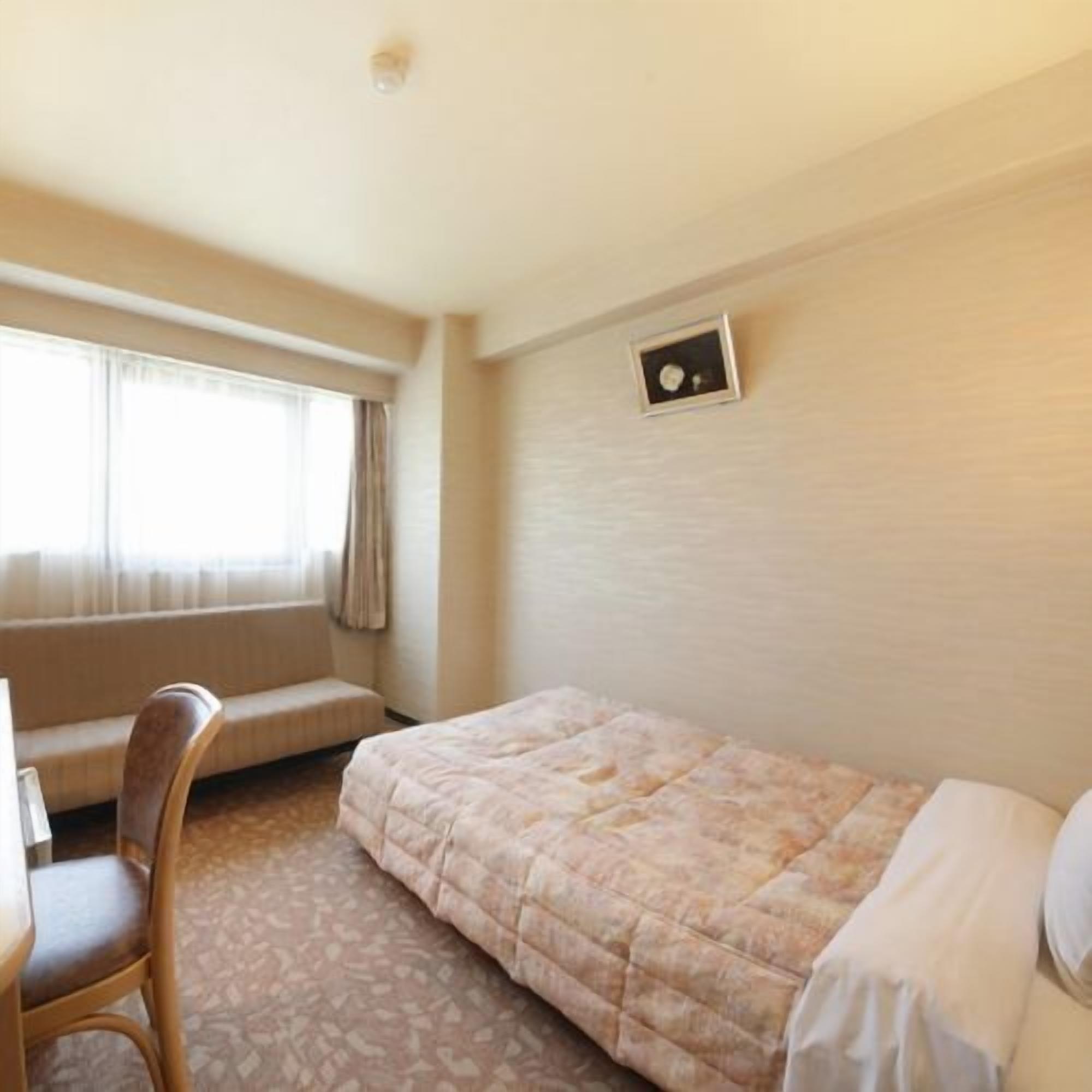 A small, simple room with a bed, a chair, a desk, and a window.