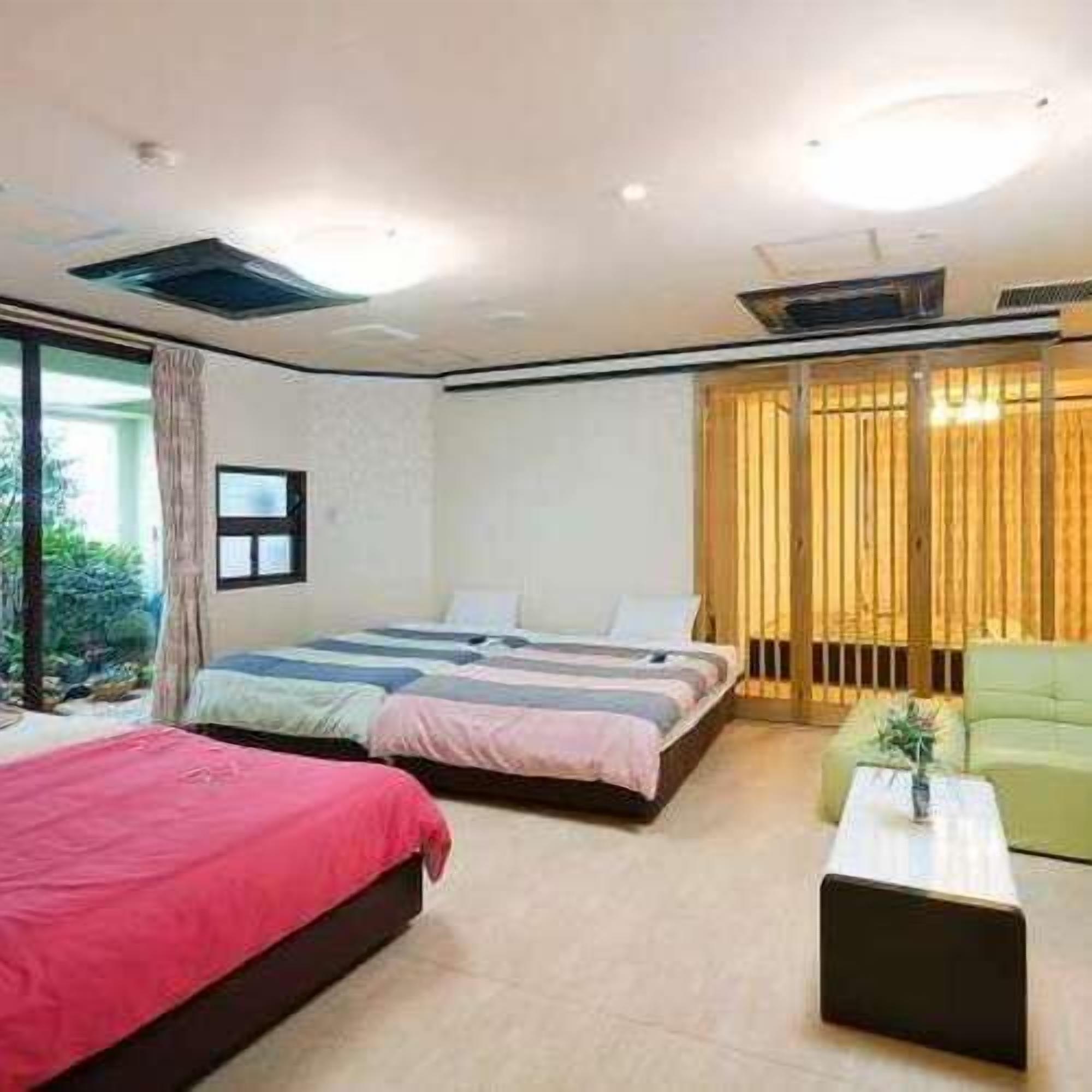 A bedroom with two beds, a green armchair, a small table, and a window with curtains.